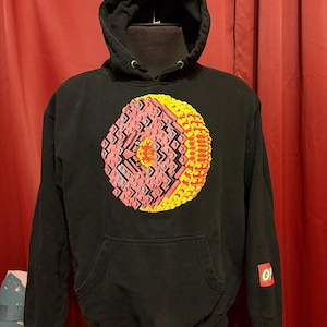 Può includere: Felpa con cappuccio nera con una grande grafica colorata sul davanti. La grafica è un design circolare con elementi rosa, gialli e blu. Un piccolo logo è sulla manica.