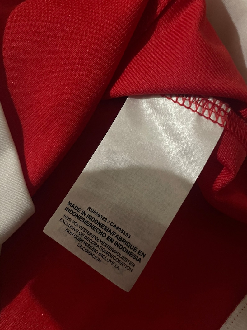 May include: A white tag with black text on a red background. The tag reads "Made in Indonesia/Fabrique en Indonesie/Hecho en Indonesia", "100% Polyester/Polyester/Poliester", "Exclusive of Decoration/Decoration Non Comprise/No Incluye La Decoracion", "RN#56323/CA#05553".