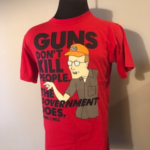 May include: Red t-shirt with a cartoon image of a man wearing glasses and a hat. The text on the shirt reads "Guns Don't Kill People. The Government Does." and "King of the Hill".