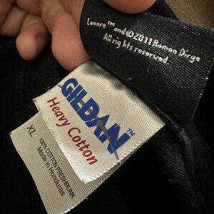 May include: Close-up of black fabric with two attached tags. The top tag reads "Lenore TM and &copy;2011 Roman Dirge All rights reserved." The bottom tag is white with "GILDAN Heavy Cotton" and "XL 100% COTTON PRESHRUNK Made in Honduras."