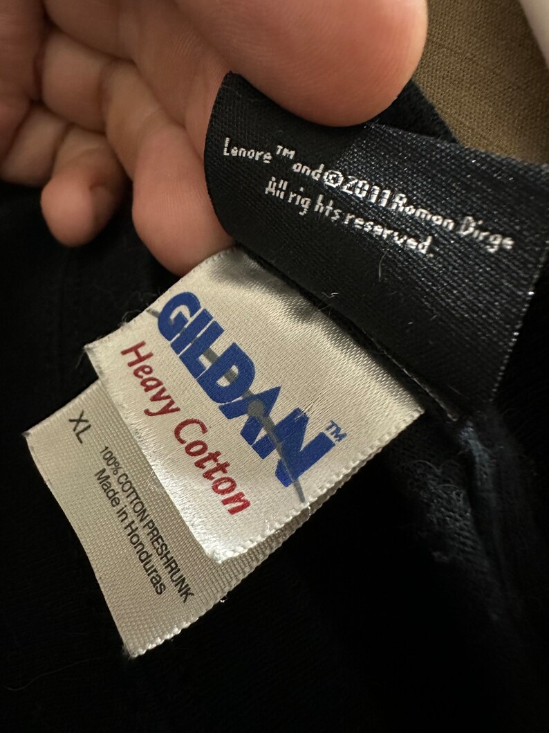 May include: Close-up of black fabric with two attached tags. The top tag reads "Lenore TM and &copy;2011 Roman Dirge All rights reserved." The bottom tag is white with "GILDAN Heavy Cotton" and "XL 100% COTTON PRESHRUNK Made in Honduras."