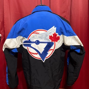 Peut inclure: Veste de baseball bleue, noire et blanche avec un grand logo au dos. Le logo représente un oiseau stylisé, une feuille d'érable et la lettre "V" à l'intérieur d'une balle de baseball. La veste a un bas et des manches noirs.