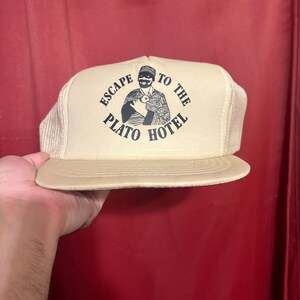 Può includere: Un cappellino trucker beige con retro in rete e pannello frontale rigido. La parte anteriore presenta una stampa grafica nera di una persona con cappello e abiti a righe, circondata dal testo circolare "ESCAPE TO THE PLATO HOTEL".