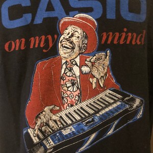 May include: A black t-shirt featuring a blue and white graphic of a man playing a keyboard. The text "CASIO on my mind" is printed on the shirt.