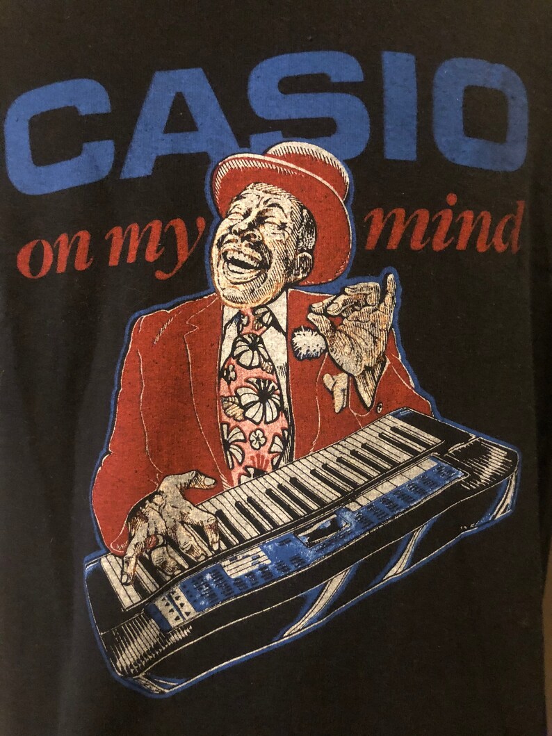 May include: A black t-shirt featuring a blue and white graphic of a man playing a keyboard. The text "CASIO on my mind" is printed on the shirt.