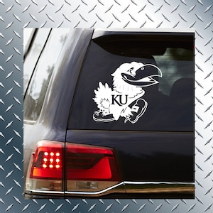 May include: White vinyl decal of a cartoon Jayhawk mascot with the letters "KU" on a car window. The Jayhawk is depicted in a running pose, with its wings spread and a determined expression. The decal is applied to a dark-colored vehicle.