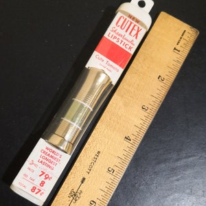 May include: A vintage CutEx Sheer Lanolin lipstick tube in a clear plastic case with a gold-coloured lipstick bullet. The lipstick is labelled "Cute Tomata" and is priced at 79 pence. The tube is sitting on a wooden ruler.