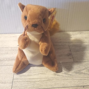 May include: A plush brown squirrel toy with a white belly, holding a small brown acorn. The squirrel has a fluffy tail and small black eyes. A tag is attached to the ear.
