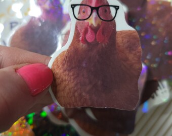 May include: A brown chicken wearing black framed glasses with a red comb and wattle. The chicken is looking directly at the camera. The image is a sticker.