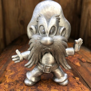 Può includere: Statuetta color argento di un personaggio dei cartoni animati con lunga barba, baffi e cappello. La figura è in posizione eretta con le braccia tese. La statuetta è su una superficie marrone arrugginita.