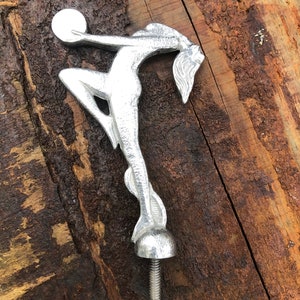 Op de afbeelding: Zilverkleurige metalen sculptuur van een vrouw die een bol vasthoudt. Het beeld is bevestigd aan een schroefdraadstang.