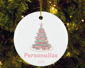 Peut inclure: Une décoration ronde en céramique blanche ornée d'une pile de livres disposés en forme de sapin de Noël, surmontée d'une étoile rose. Le mot "Personalize" est imprimé en rose sous l'arbre, avec l'année "2026" centrée en dessous.