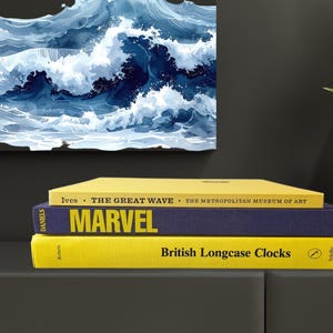 May include: A stack of three hardcover books with yellow and navy blue covers. The top book is titled "Ives The Great Wave The Metropolitan Museum of Art". The middle book says "MARVEL". The bottom book says "British Longcase Clocks".