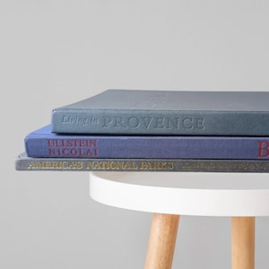 May include: A stack of three hardcover books. The top book is blue with the text Living in PROVENCE. The middle book is purple with the text ULLSTEIN NICOLAI BERLIN. The bottom book is dark gray with the text AMERICA'S NATIONAL PARKS.