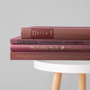 May include: A stack of four hardcover books in shades of maroon and burgundy. The spines feature gold and black text including titles like Motorcycles: An Illustrated History, The Jubilee Book, and Mirror of the Invisible World, resting on a white stool.
