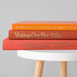 May include: A stack of three hardcover books resting on a white stool. The top book is bright orange with the text Barefoot Contessa Family Style. The middle book is rust-colored with the text Visiting Our Past America's Historylands. The bottom book is red.