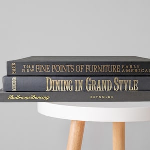 May include: A stack of three hardcover books with dark gray covers and gold lettering. The titles are The New Fine Points of Furniture Early American, Dining in Grand Style, and Ballroom Dancing, with author names Sack, Schneider, and Reynolds visible.