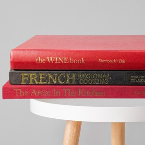 May include: A stack of three hardcover books on a white table. The top book is red with "the WINE book" in gold lettering. The middle book is black with "FRENCH REGIONAL COOKING" in gold. The bottom book is red with "The Artist In The Kitchen".