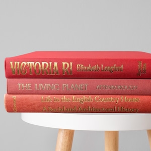 May include: Three hardcover books stacked on a white table. The top book is red with gold lettering that reads "VICTORIA RI". The middle book is pink and reads "THE LIVING PLANET". The bottom book is orange and reads "Life in the English Country House".