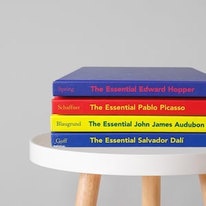 May include: A stack of four books with colorful covers, each titled "The Essential" followed by an artist's name: Edward Hopper, Pablo Picasso, John James Audubon, and Salvador Dalí. The books are on a white table.