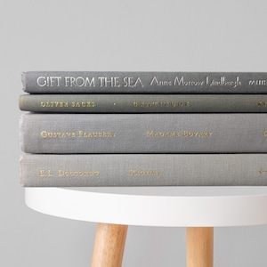 May include: A stack of four hardcover books with gray fabric covers and gold foil lettering on the spines. The titles include Gift from the Sea by Anne Morrow Lindbergh, Gratitude by Oliver Sacks, Madame Bovary by Gustave Flaubert, and Ragtime by E.L. Doctorow.