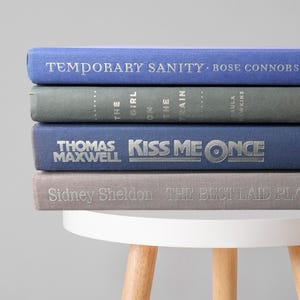 May include: A stack of four hardcover books with titles and author names in silver lettering. The books are in shades of blue and gray. The titles include "Temporary Sanity", "Kiss Me Once", and "The Best Laid Plans".