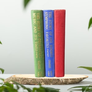 May include: Three hardcover books with gold lettering on a wooden stand. The books are titled "The Story of One Hundred Symphonic Favorites", "The Story of Orchestral Music and Its Times", and "The Story of Orchestral Music and Its Times".