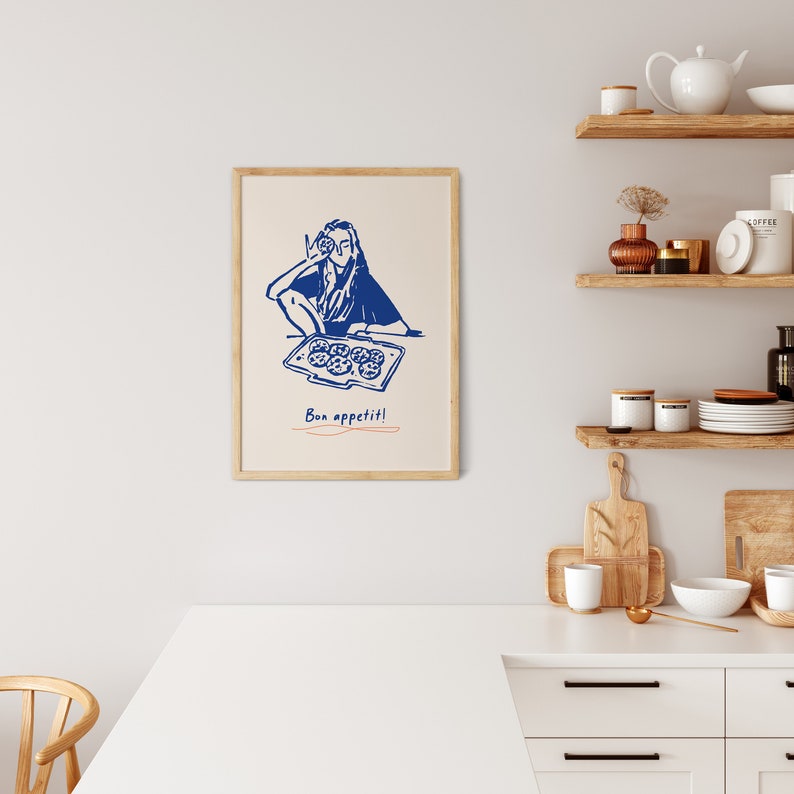May include: A blue line drawing of a person holding a tray of cookies with the text "Bon app&eacute;tit!" underneath. The drawing is framed in a light wood frame and is hanging on a white wall.