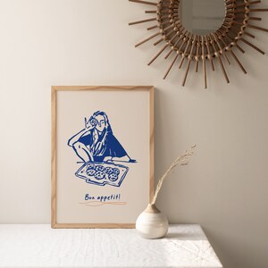 May include: A framed illustration of a person holding a cookie in front of their face, with a baking sheet of cookies in front of them. The illustration is in blue on a white background. The text "Bon appetit!" is written below the illustration.