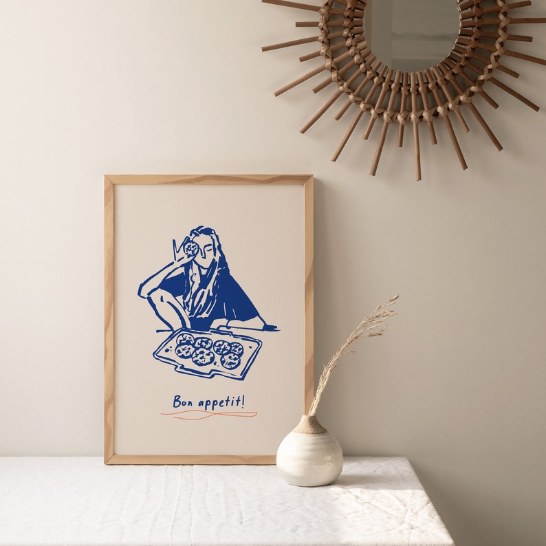May include: A framed illustration of a person holding a cookie in front of their face, with a baking sheet of cookies in front of them. The illustration is in blue on a white background. The text "Bon appetit!" is written below the illustration.