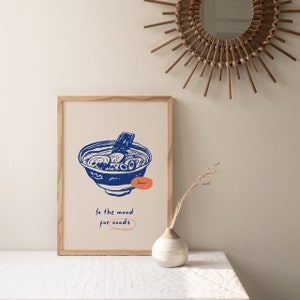 May include: A framed print with a blue illustration of a bowl of ramen noodles. The text "Ramen" is written in orange below the bowl. The text "In the mood for noodles" is written in blue below the bowl.