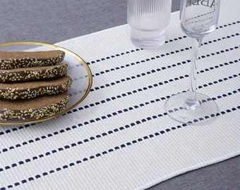 May include: White table runner with navy blue dotted lines. The runner is on a gray surface with a glass of water and a glass of wine on top of it. There is a plate of bread on the runner.