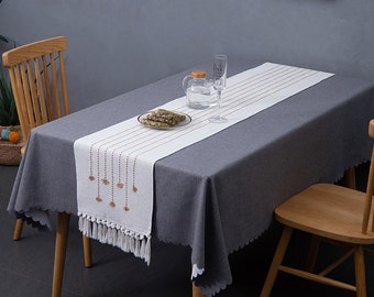 May include: White table runner with a fringe border and a design of brown tassels hanging from a string of beads. The runner is on a gray tablecloth on a wooden table.
