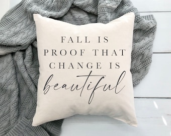 Puede incluir: Una almohada blanca con el texto "Fall is proof that change is beautiful" en letras negras. La almohada está sobre una manta de punto gris.