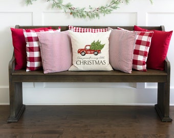 May include: A wooden bench is adorned with red and white Christmas-themed pillows. The pillows feature solid red, red and white gingham, and red and white striped patterns. A central pillow displays a red truck carrying a Christmas tree, with the text "Have yourself a very little CHRISTMAS."