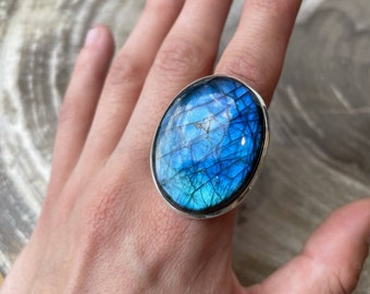 May include: A silver ring with a large oval blue labradorite gemstone. The stone has a iridescent sheen and a unique pattern.