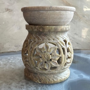 Puede incluir: Un difusor de aceite de piedra tallada con una parte superior en forma de cuenco y una base decorativa. La base presenta intrincados recortes florales y geométricos. El color es un verde-gris apagado, lo que le da un aspecto natural y terroso. El difusor está diseñado para contener aceites esenciales.