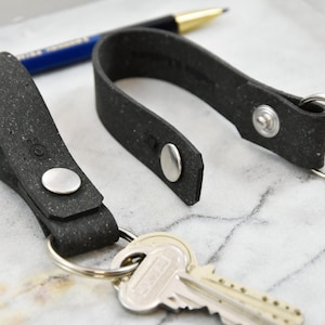 May include: Two black leather keychains with silver metal rings and snaps. The keychains are made of a textured material that resembles recycled paper. One keychain has a key attached to it.