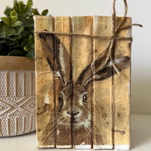 May include: A stack of vintage books with a painted image of a rabbit's face on the front. The books are tied together with twine and have a rustic, antique look.