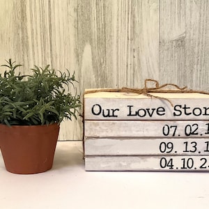 May include: A stack of white books with the text "Our Love Story" and dates "07.02.15", "09.13.19", and "04.10.22" printed on the covers. The books are tied together with twine.