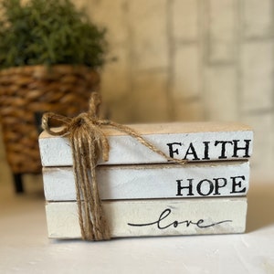 May include: A stack of three white painted books with the words "FAITH", "HOPE", and "love" written in black. The books are tied together with twine.