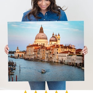 May include: A framed canvas print of Venice, Italy, featuring the Grand Canal with buildings, domes, and a boat. The print has a blue sky and water, with warm-toned buildings. The canvas is held by a person.
