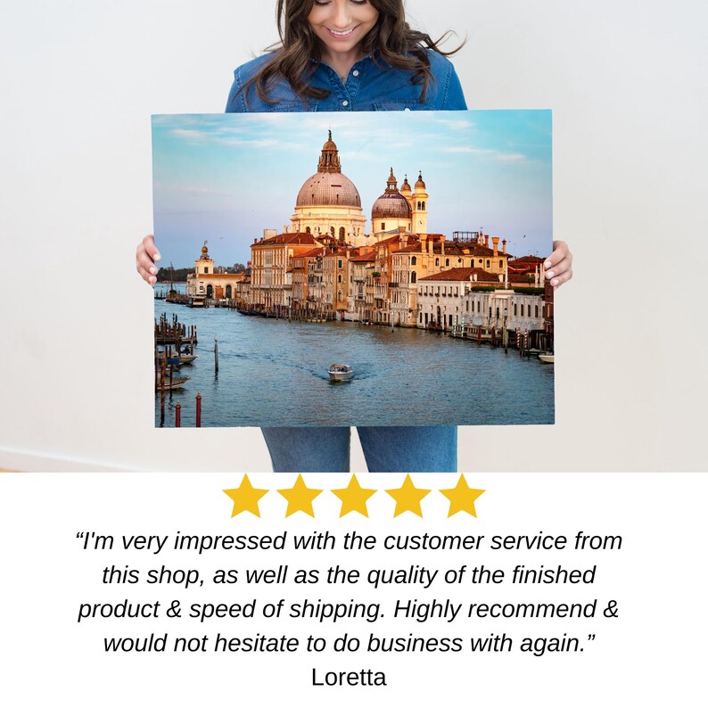 May include: A canvas print of a scenic view of Venice, Italy, featuring the Grand Canal, buildings, and a boat. The print has a blue sky and water, with a warm color palette. The print is held by a person.
