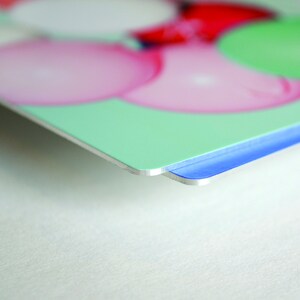 May include: Close-up of a stack of rectangular, colorful art prints. The top print is light green, the middle print is blue, and the background shows blurred pink and green shapes. The edges of the prints are silver.