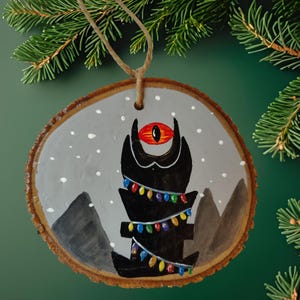 May include: Hand-painted wooden Christmas ornament. The round ornament features a black creature with a red eye, decorated with colorful lights. The background is gray with white snow dots and gray mountains.