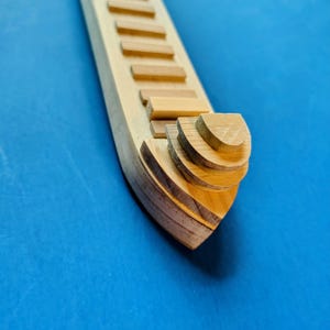 Może przedstawiać: Drewniany przedmiot w kształcie łodzi z szeregiem prostokątnych stopni wzdłuż jego długości. Przód łodzi ma warstwową, zakrzywioną konstrukcję. Drewno ma naturalny, jasnobrązowy kolor na niebieskim tle.