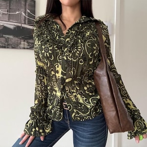 Peut inclure: Chemisier à manches longues avec un motif floral vert et jaune. Le chemisier a un col et des poignets à volants. Un sac à bandoulière en cuir marron est également visible.