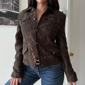 May include: A dark brown paisley-patterned jacket with a button-up front, two chest pockets, and two lower pockets. The jacket has a collared neckline and gold-colored buttons. The jacket is worn with blue jeans.