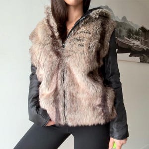May include: A hooded jacket with a faux fur front and hood, and black sleeves. The faux fur is a mix of brown and tan tones. The jacket has a zip closure.