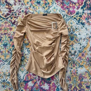 Peut inclure: Un haut à manches longues de couleur beige avec des détails froncés et un col carré. Le haut présente une boucle décorative argentée sur le devant et des manches froncées. Le haut est fait d'un tissu doux et fluide.